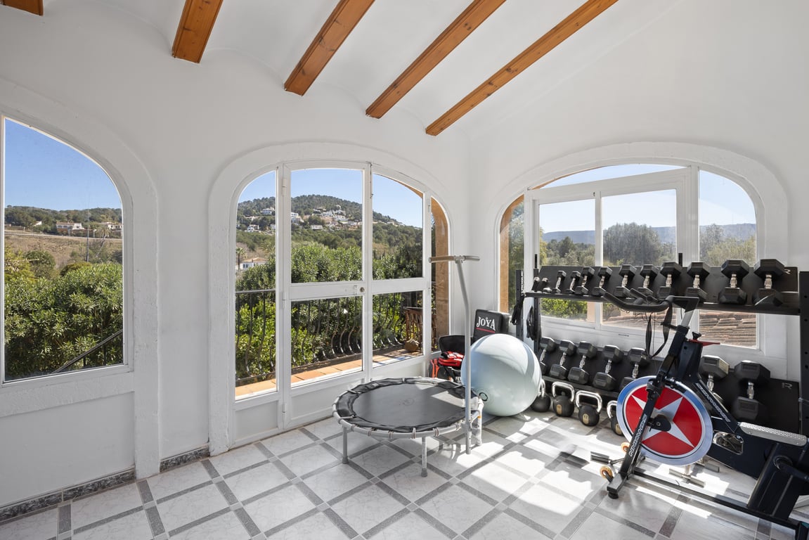 Naya vitrée aménagée en salle de sport, dotée de poutres en bois, d'un sol en grès et de grandes arches offrant une vue dégagée sur les collines.