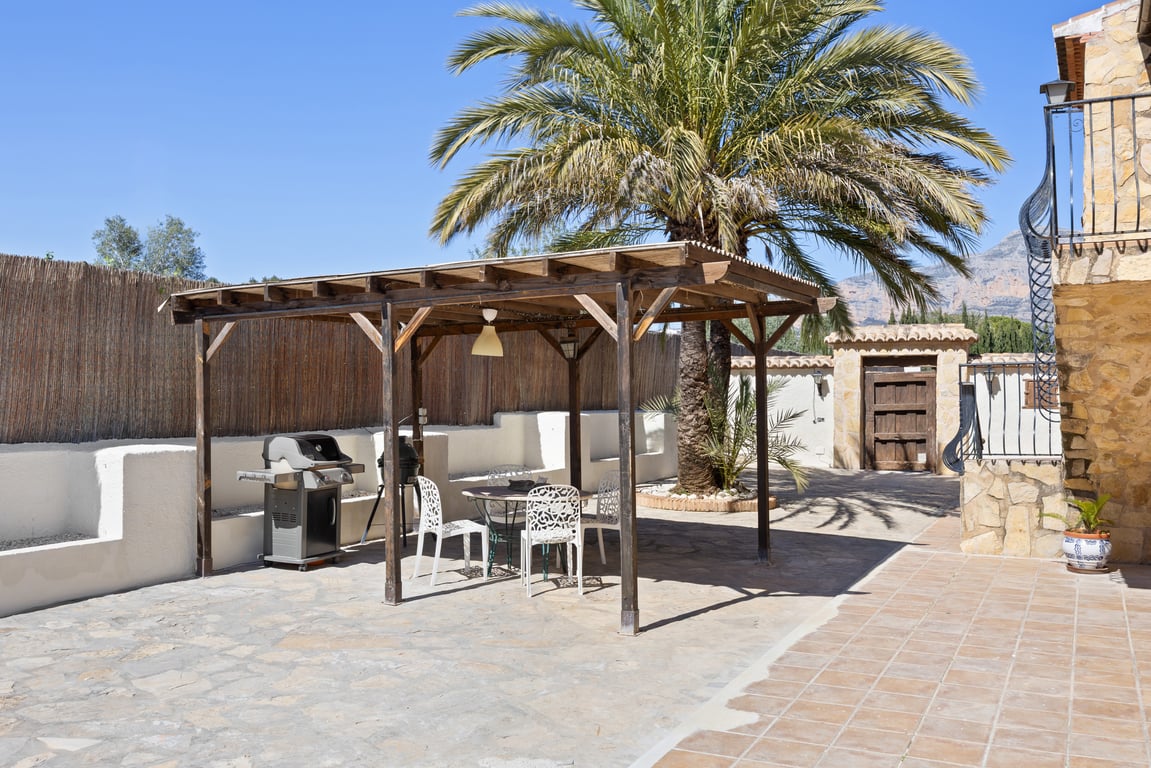 Pergola en bois abritant un coin repas extérieur avec barbecue, sur une terrasse pavée agrémentée d'un grand palmier et de murs en pierre.