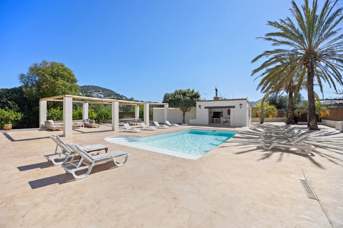 Piscine de 5x10m entourée d'une vaste terrasse pavée, comprenant une cuisine d'été équipée, une pergola en bois et des palmiers méditerranéens.