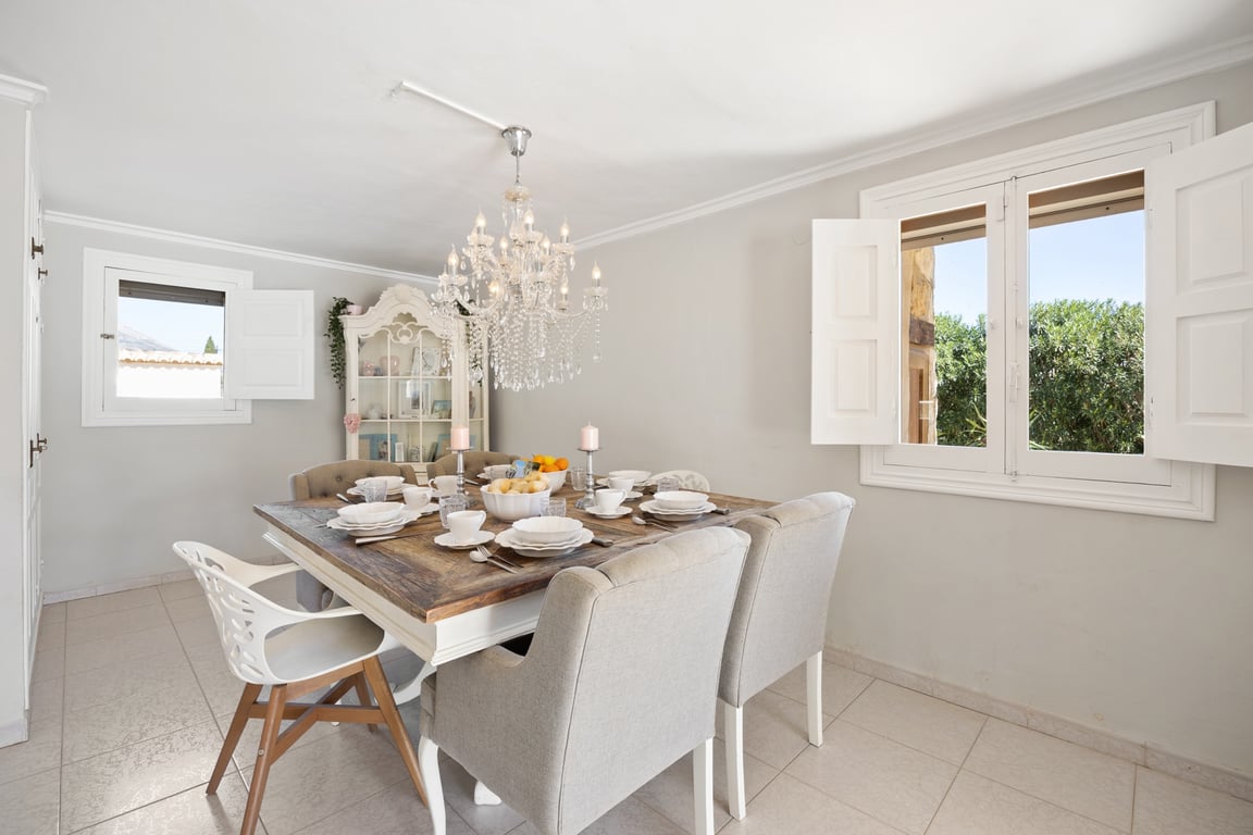 Salle à manger lumineuse avec table en bois rustique, chaises rembourrées et lustre en cristal. Les fenêtres à volets blancs offrent une vue dégagée sur l'extérieur.