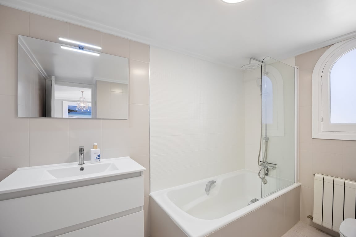 Salle de bain moderne avec meuble vasque blanc, grand miroir éclairé et baignoire avec paroi en verre. Une fenêtre en arc apporte de la lumière naturelle aux tons neutres.