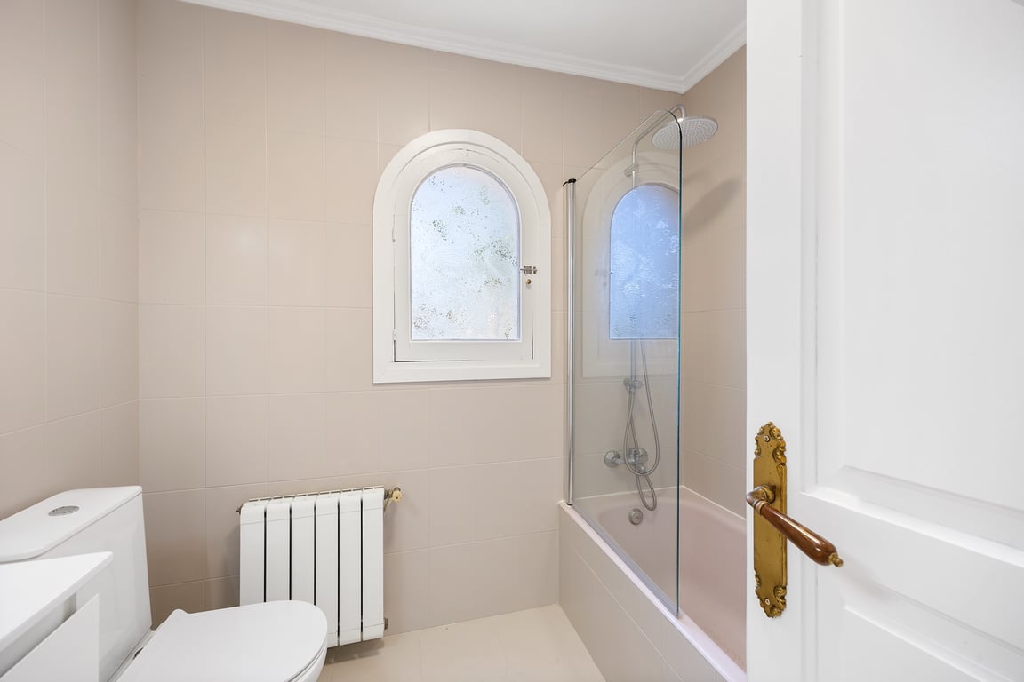 Salle de bains fonctionnelle avec baignoire, paroi en verre et fenêtre en plein cintre. Équipée d'un radiateur et de finitions dans des tons neutres.