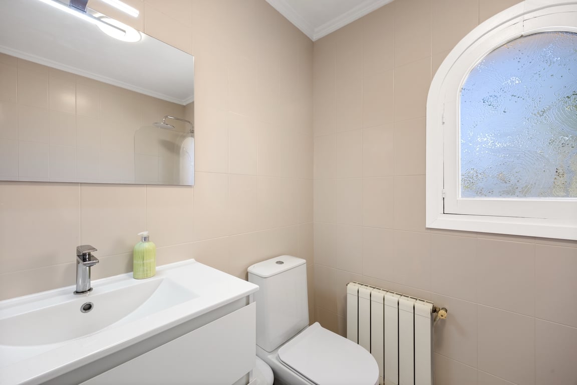 Salle de bains fonctionnelle avec carrelage neutre, meuble vasque blanc, radiateur et une fenêtre cintrée avec verre dépoli apportant une luminosité douce.