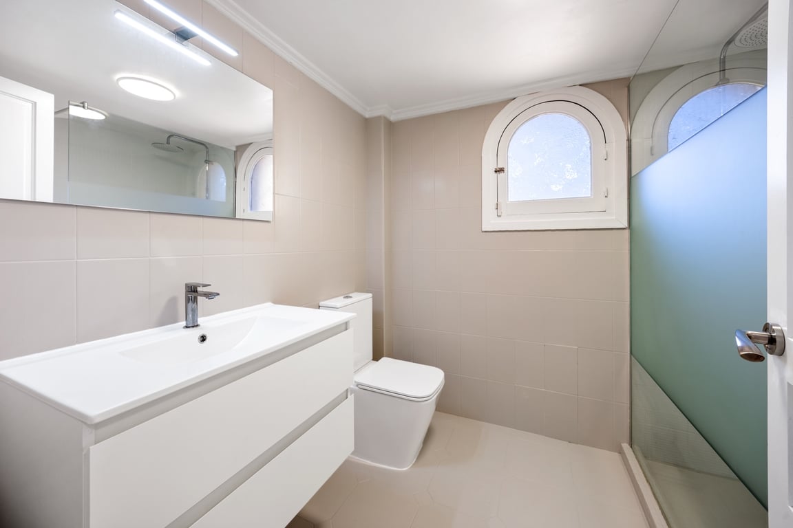 Salle de bains moderne avec meuble vasque blanc, douche à l'italienne et fenêtre en arc. Carrelage beige élégant et éclairage LED intégré au-dessus du miroir.