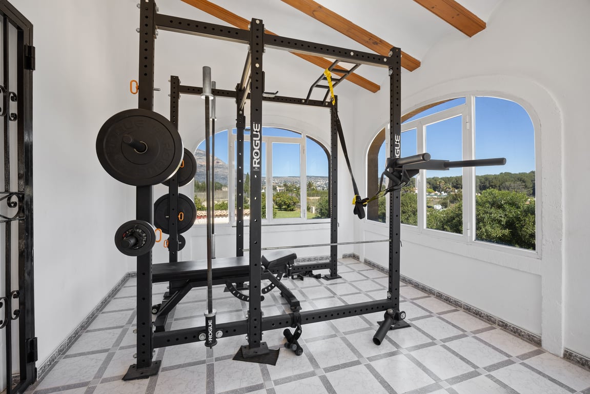 Salle de sport privée équipée d'un rack Rogue professionnel, située dans une pièce lumineuse avec poutres en bois et vues dégagées sur la nature environnante.