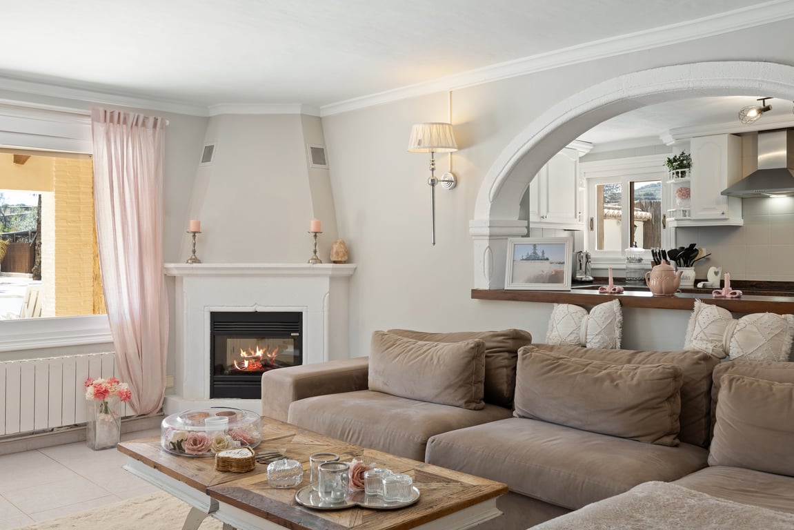 Salon lumineux avec cheminée d'angle, canapé modulaire aux tons neutres et une arche blanche ouvrant sur la cuisine équipée.