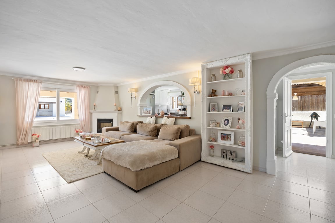 Salon lumineux avec cheminée d'angle, canapé modulaire et ouvertures en arche vers la cuisine et l'extérieur. Comprend une bibliothèque décorative et un sol carrelé.