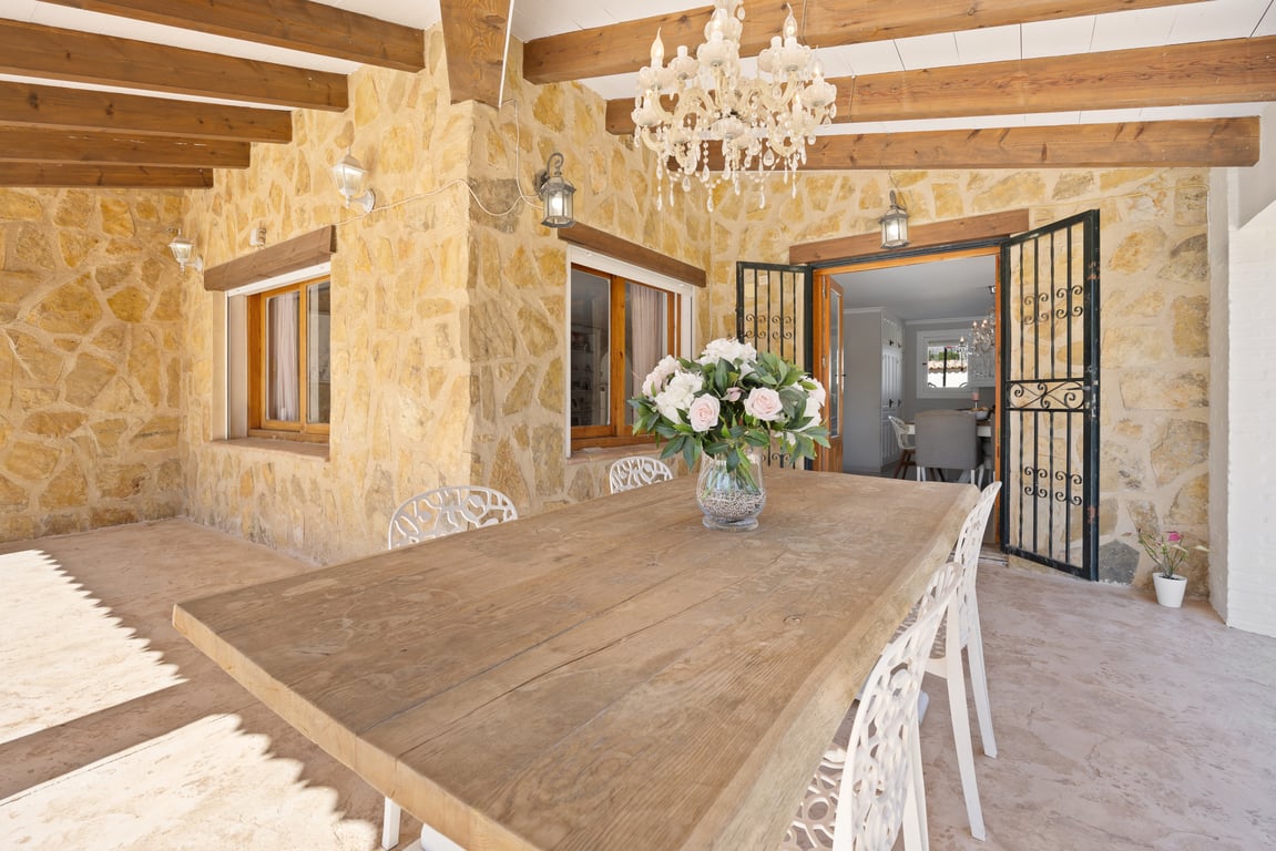 Terrasse couverte avec murs en pierre naturelle, poutres en bois apparentes et une grande table à manger sous un lustre élégant.