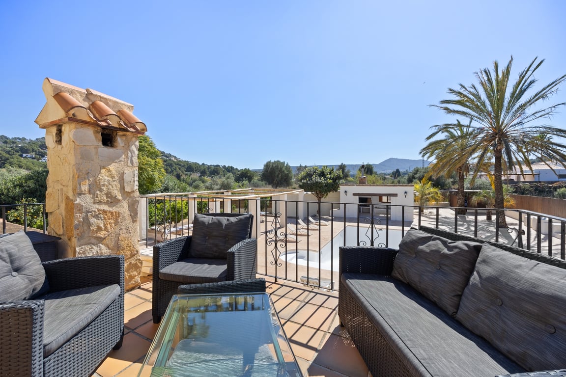 Terrasse supérieure avec salon de jardin en rotin, sols en terre cuite et vue sur la piscine, la cuisine d'été et les montagnes environnantes.