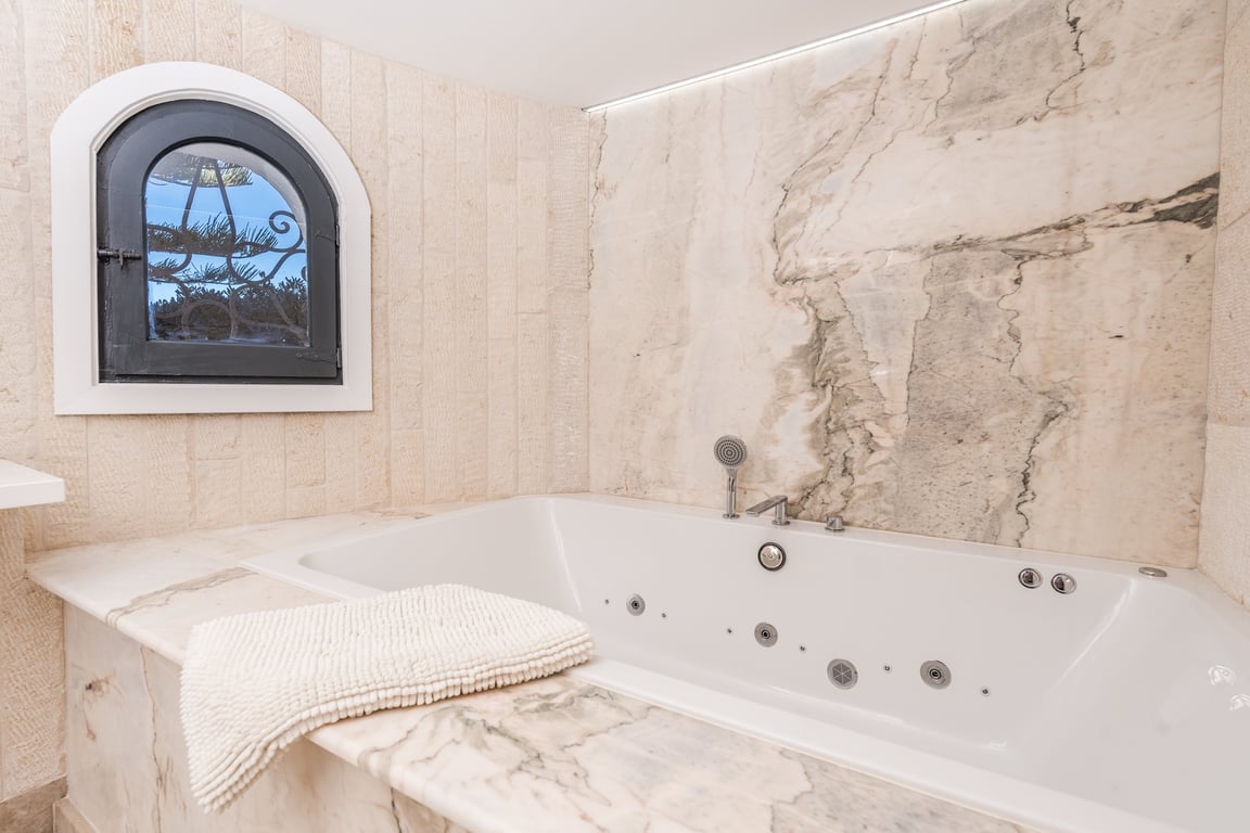 Baignoire d'hydromassage encastrée avec finitions en marbre veiné, murs en pierre naturelle et une fenêtre cintrée authentique.