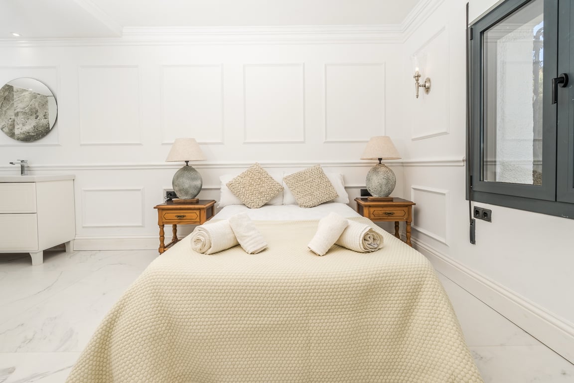 Chambre avec boiseries blanches et sol en marbre. Elle comprend un coin vasque intégré avec miroir circulaire et des tables de chevet en bois classique.