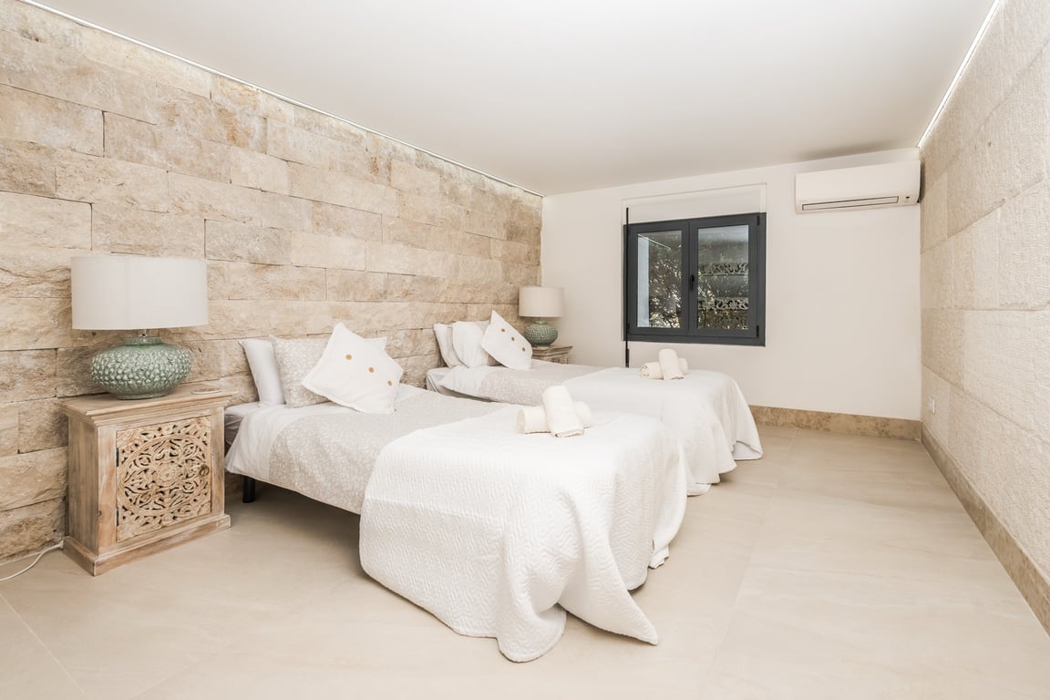 Chambre avec deux lits simples, un mur en pierre naturelle, des tables de chevet en bois sculpté et un éclairage LED discret au plafond.
