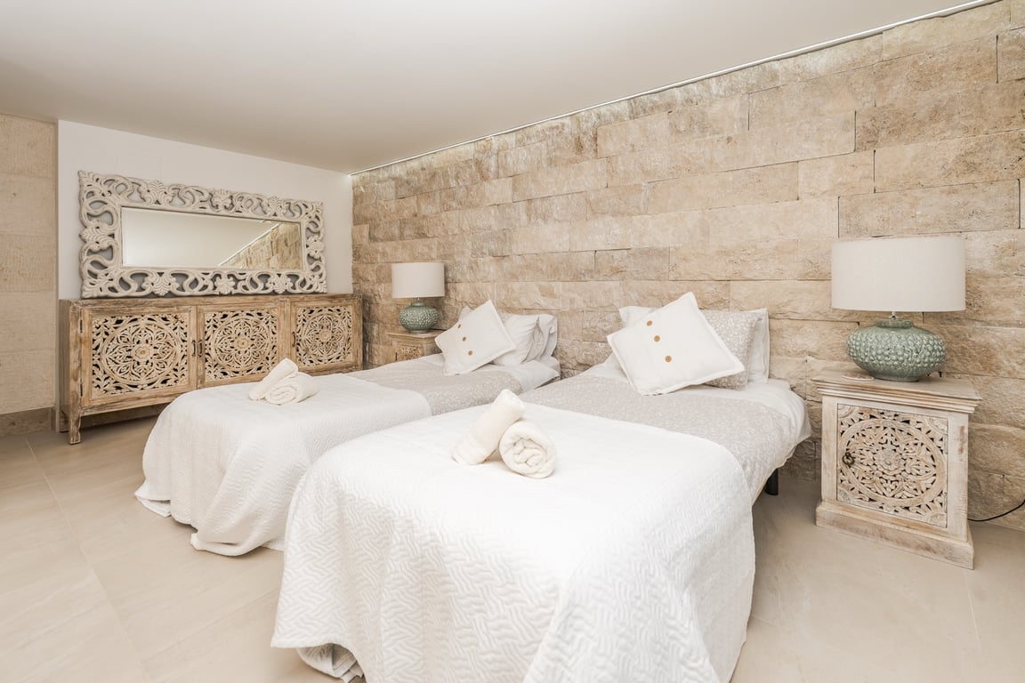 Chambre avec lits jumeaux, mur en pierre naturelle apparente et mobilier en bois sculpté. Éclairage encastré au plafond et ambiance méditerranéenne aux tons sereins.