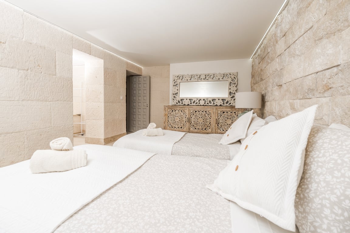 Chambre avec murs en blocs de pierre calcaire, lits jumeaux et mobilier en bois sculpté. Un éclairage LED indirect souligne les textures naturelles du mur.
