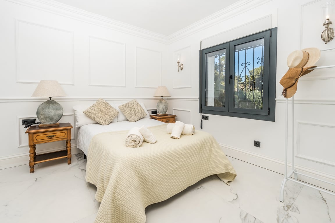 Chambre lumineuse avec moulures murales décoratives, sol en marbre blanc et fenêtre à cadre sombre. Équipée de tables de chevet en bois et de lampes en pierre.