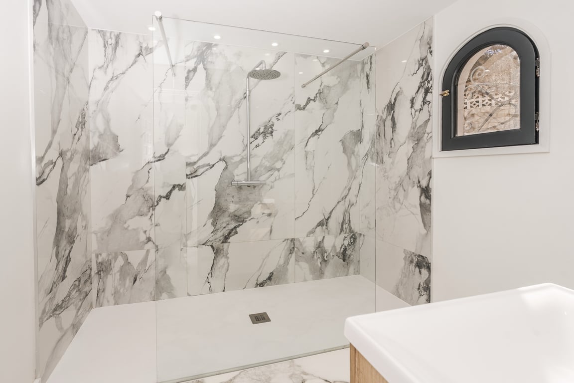 Douche à l'italienne avec parois en marbre, paroi vitrée fixe et fenêtre cintrée à cadre noir, alliant design contemporain et détails architecturaux classiques.