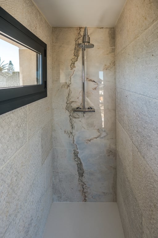Douche à l'italienne avec un mur en marbre veiné et des parois latérales en pierre calcaire texturée. Comprend une robinetterie chromée et une fenêtre latérale.