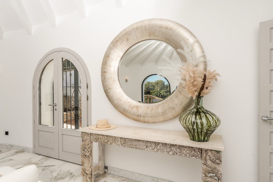 Entrée avec sol en marbre, console en bois sculpté et grand miroir circulaire. Les plafonds à poutres blanches et les portes cintrées soulignent le design méditerranéen.