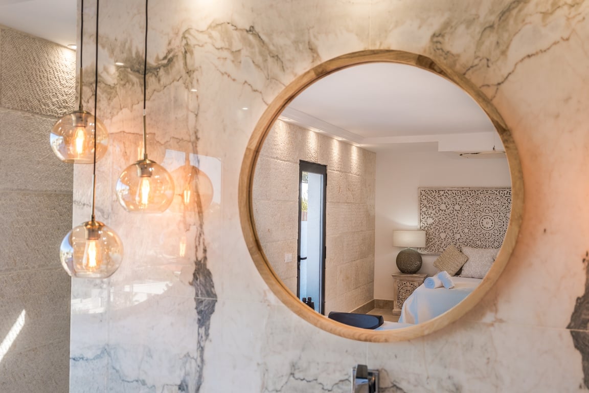 Miroir circulaire avec cadre en bois sur un mur en marbre veiné, éclairé par des suspensions en verre et reflétant une chambre aux murs en pierre caliza.