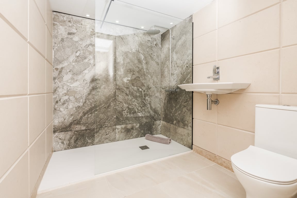 Salle de bain moderne avec douche à l'italienne, paroi en verre et mur en marbre gris. Équipée de sanitaires blancs et de carrelage aux tons crème.