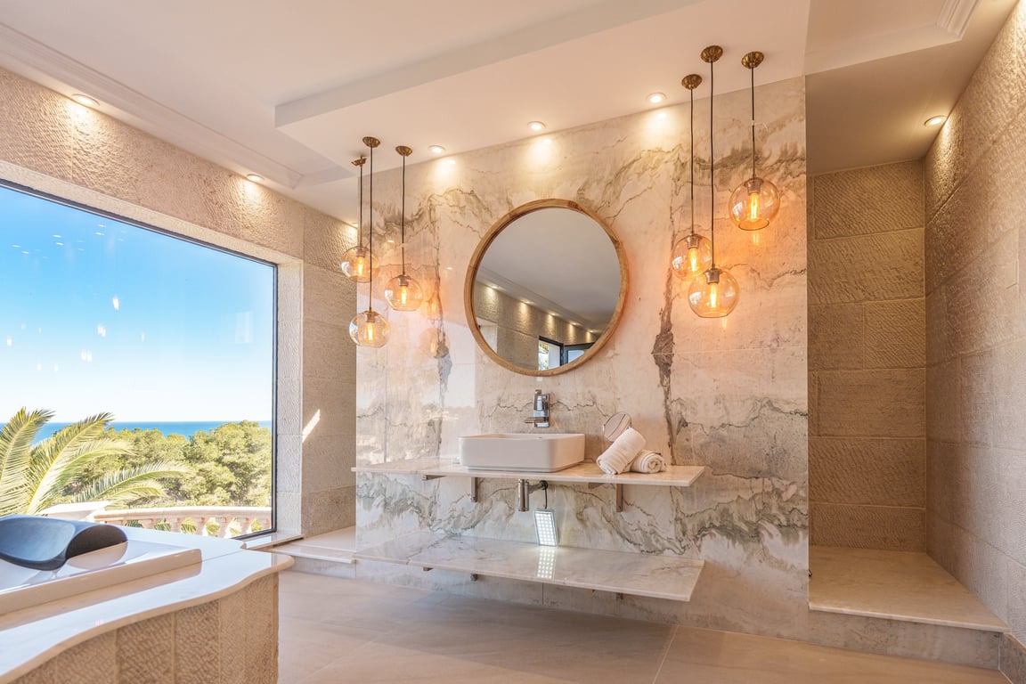 Salle de bain principale avec murs en marbre veiné, vasque à poser sur plan suspendu et miroir circulaire. Des suspensions en verre et une large fenêtre ouvrent sur la mer et la côte.