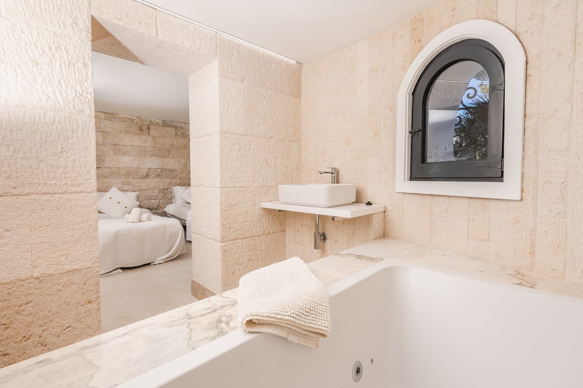 Salle de bains attenante avec murs en pierre naturelle, baignoire bordée de marbre et vasque moderne, offrant une vue sur la chambre à travers une arche en pierre.