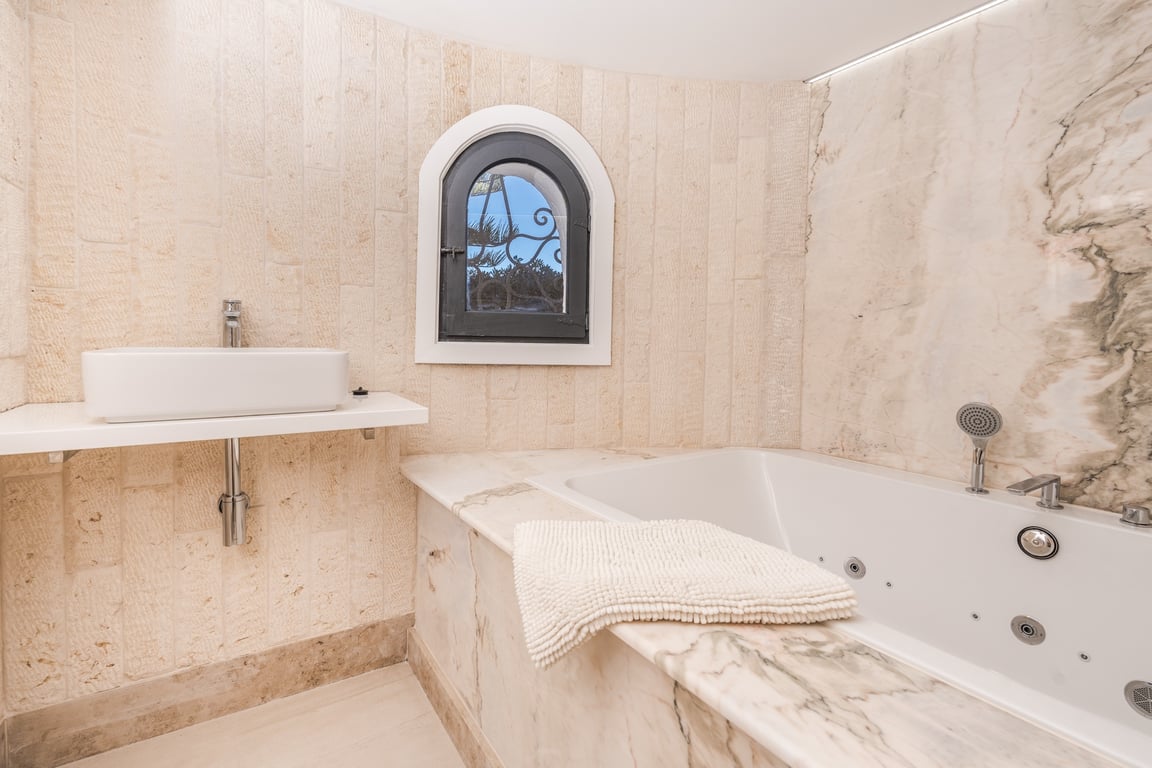 Salle de bains avec baignoire balnéo habillée de marbre, vasque à poser et murs en pierre naturelle agrémentés d'une fenêtre en plein cintre.