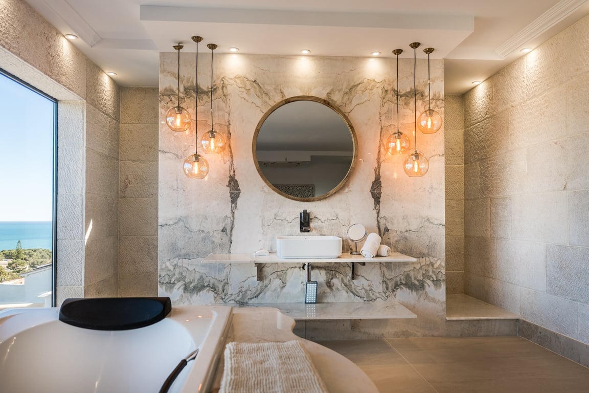 Salle de bains de la suite parentale avec mur en marbre veiné, vasque à poser sur étagère flottante, suspensions design et vue panoramique sur la mer.