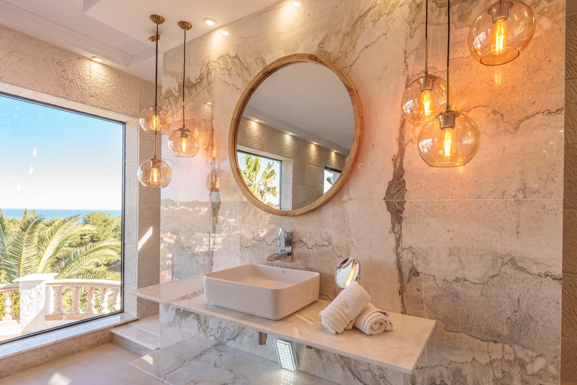 Salle de bains de la suite parentale avec murs en marbre, vasque à poser, miroir rond en bois et suspensions en verre offrant une vue sur la mer.