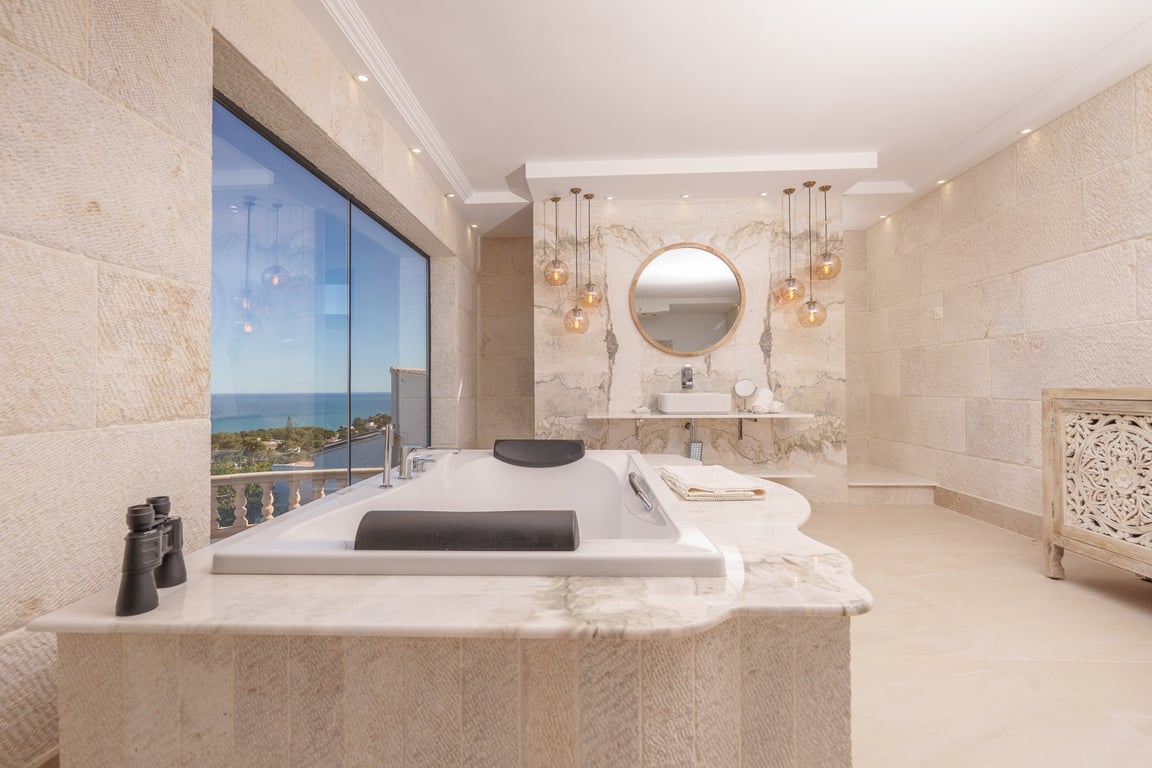 Salle de bains de la suite parentale avec murs en pierre calcaire, baignoire encastrée en marbre offrant une vue sur mer et luminaires suspendus décoratifs.
