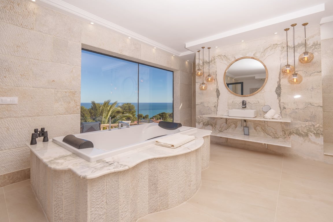 Salle de bains de la suite principale avec baignoire en marbre, murs en pierre calcaire et vue panoramique sur la mer à travers une grande baie.