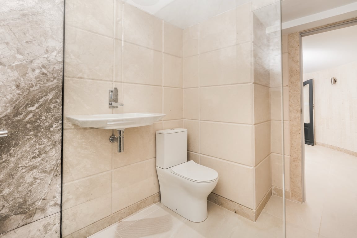 Salle d'eau moderne avec carrelage en pierre calcaire crème, toilettes contemporaines et lavabo suspendu. Présence d'une paroi en verre et de détails en marbre.