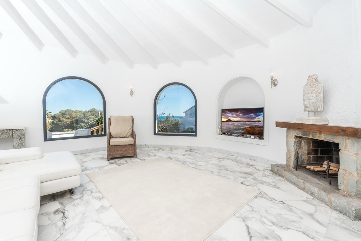 Salon circulaire avec plafond à poutres blanches, sol en marbre et cheminée en pierre rustique. Les fenêtres cintrées offrent une vue sur la mer et la végétation environnante.