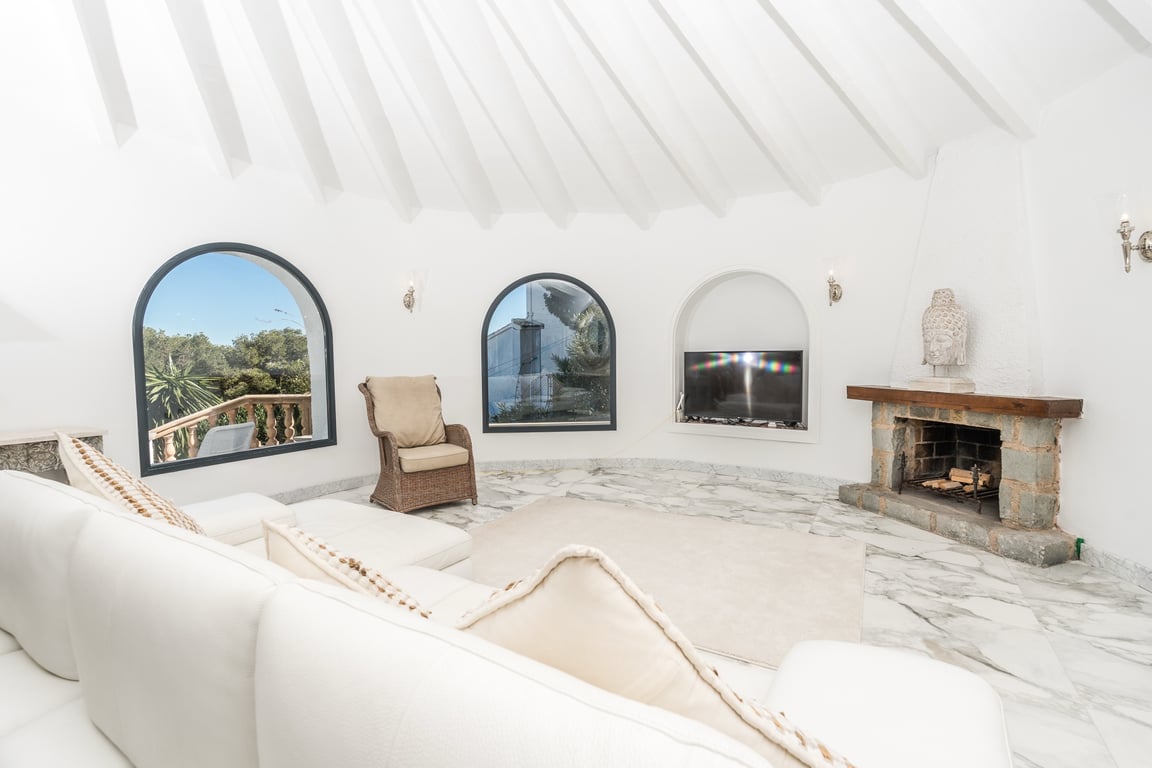 Salon circulaire avec plafond à poutres radiales blanches, sol en marbre et cheminée en pierre. Les fenêtres en arc offrent une vue dégagée sur l'extérieur.