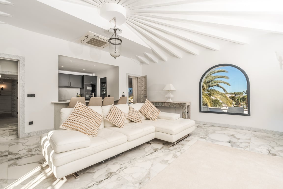 Salon lumineux avec sol en marbre, plafond à poutres radiales et canapé blanc. Une fenêtre en arche offre une vue sur les palmiers, à côté d'une cuisine semi-ouverte.
