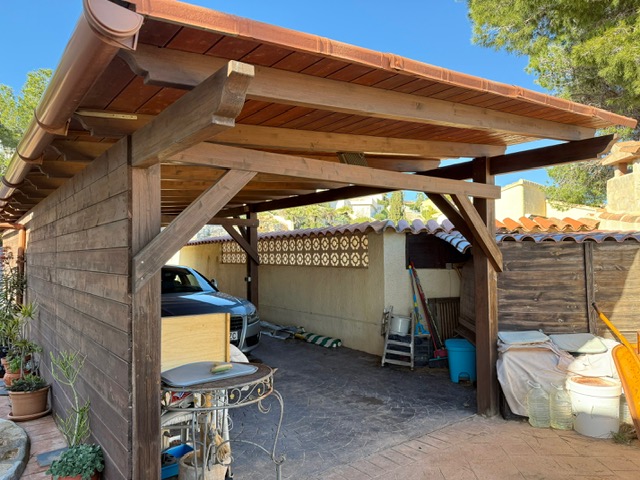 Abri de voiture en bois avec poutres apparentes et toit en tuiles, offrant un espace de stationnement couvert sur un sol en béton imprimé.