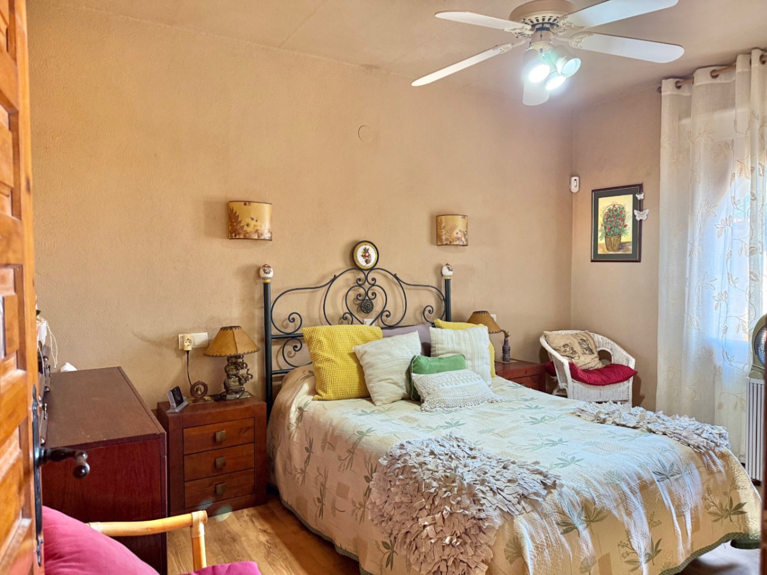 Chambre avec tête de lit en fer forgé, tables de chevet en bois et ventilateur de plafond. La pièce offre des tons chauds et un fauteuil en osier près de la fenêtre.