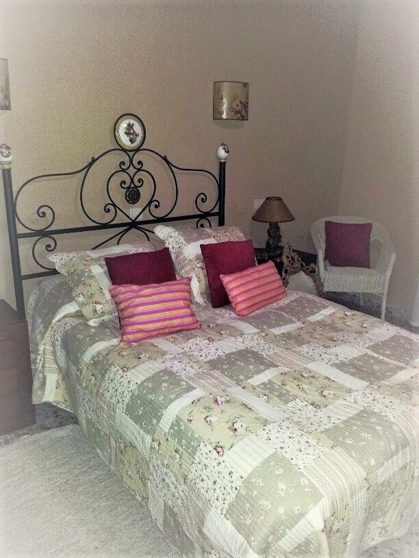 Chambre avec un lit double, une tête de lit en fer forgé noir et un couvre-lit en patchwork. Elle comprend un fauteuil en osier blanc et des murs aux tons neutres.