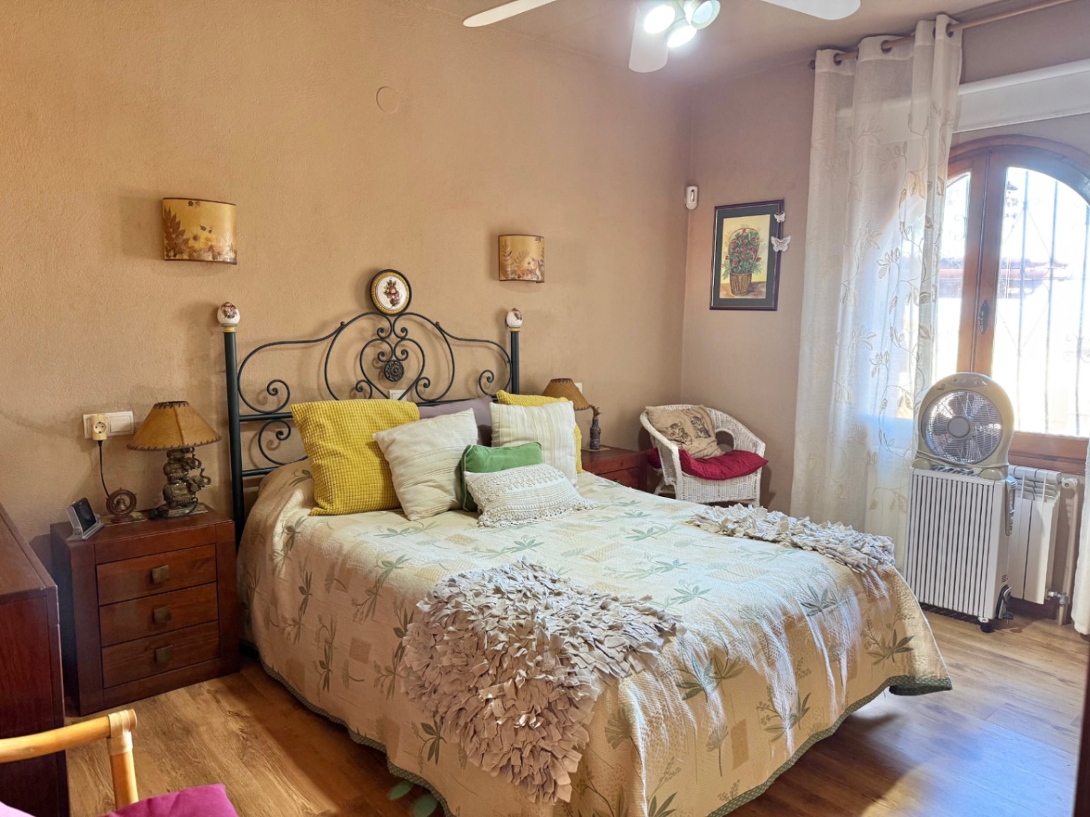 Chambre rustique avec tête de lit en fer forgé, parquet et fenêtre en arc. Comprend un ventilateur de plafond, un radiateur et des tables de chevet en bois.