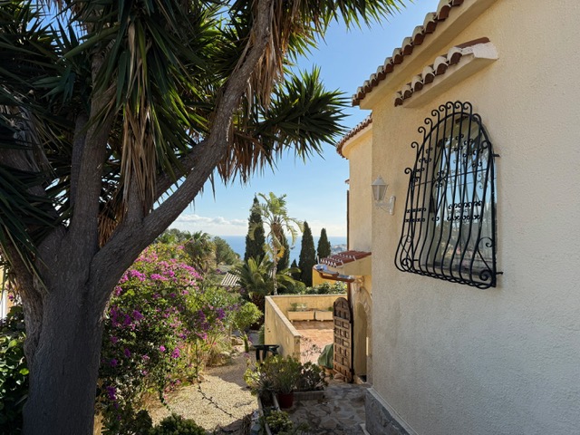 Façade extérieure avec fenêtres en fer forgé, entourée d'un jardin méditerranéen et de bougainvilliers offrant une vue sur la mer.
