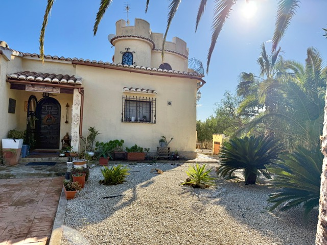 Façade principale de la villa avec sa tour circulaire distinctive, porche d'entrée couvert et jardin méditerranéen en gravier avec palmiers.