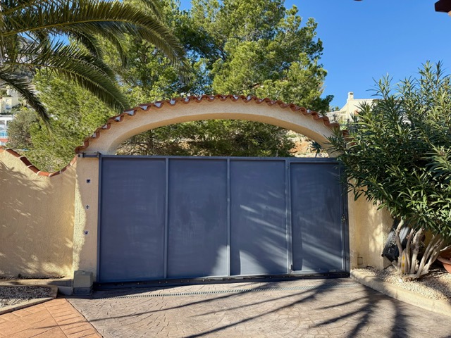 Portail d'entrée coulissant sous une arche méditerranéenne avec tuiles en terre cuite, entouré de végétation et d'une allée pavée.