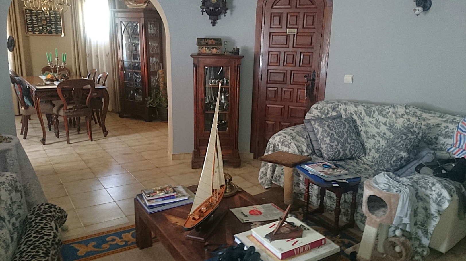 Salon traditionnel avec sol en céramique, arche ouvrant sur la salle à manger et mobilier en bois classique, comprenant des vitrines et une table basse avec décoration nautique.