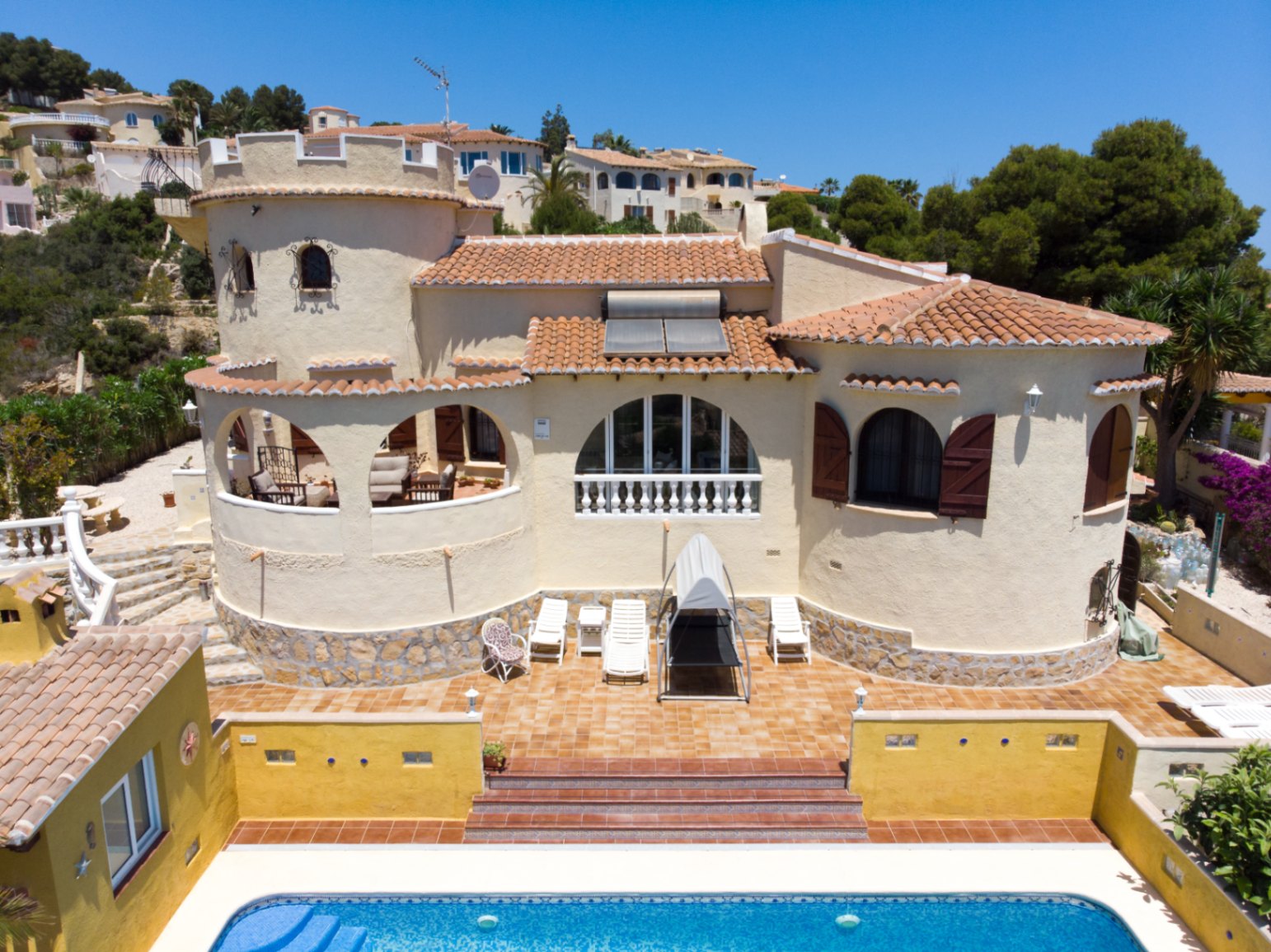 Vue extérieure de la villa avec tour circulaire, terrasses voûtées et piscine. Présente des volets en bois, un toit en tuiles avec panneaux solaires et un soubassement en pierre.