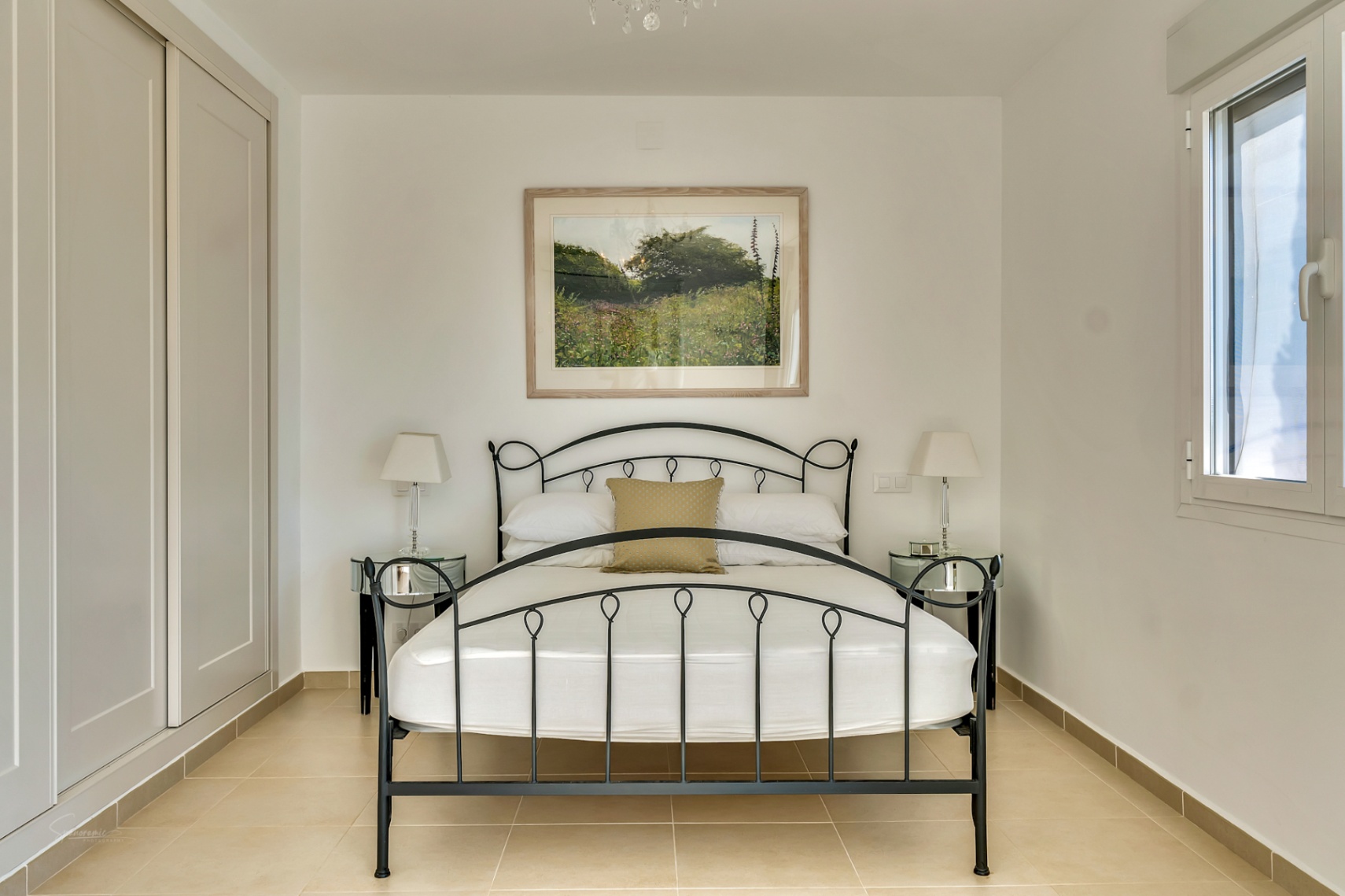 Chambre avec cadre de lit en fer forgé noir, armoires encastrées, tables de chevet en miroir et sol en carrelage clair éclairé par une fenêtre.