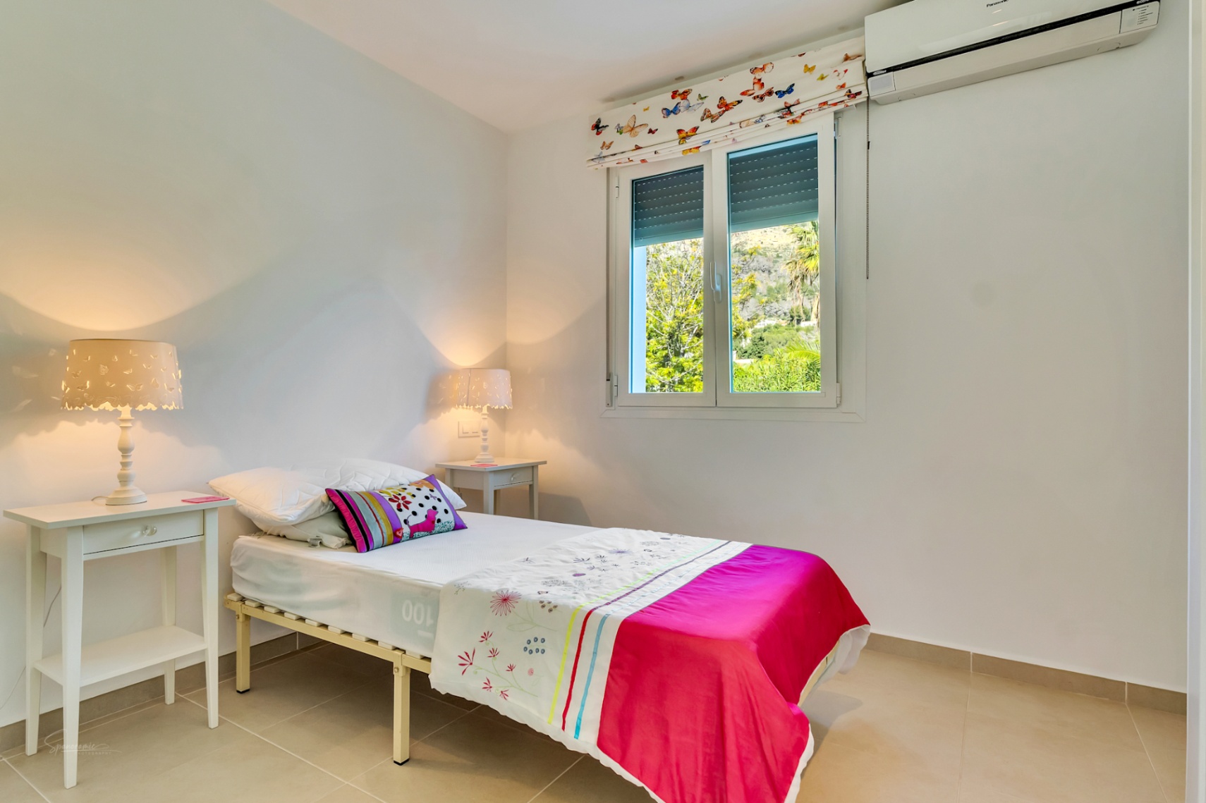 Chambre avec lit simple, climatisation split et fenêtre vue jardin. Comprend des tables de chevet blanches et des lampes aux motifs de papillons.