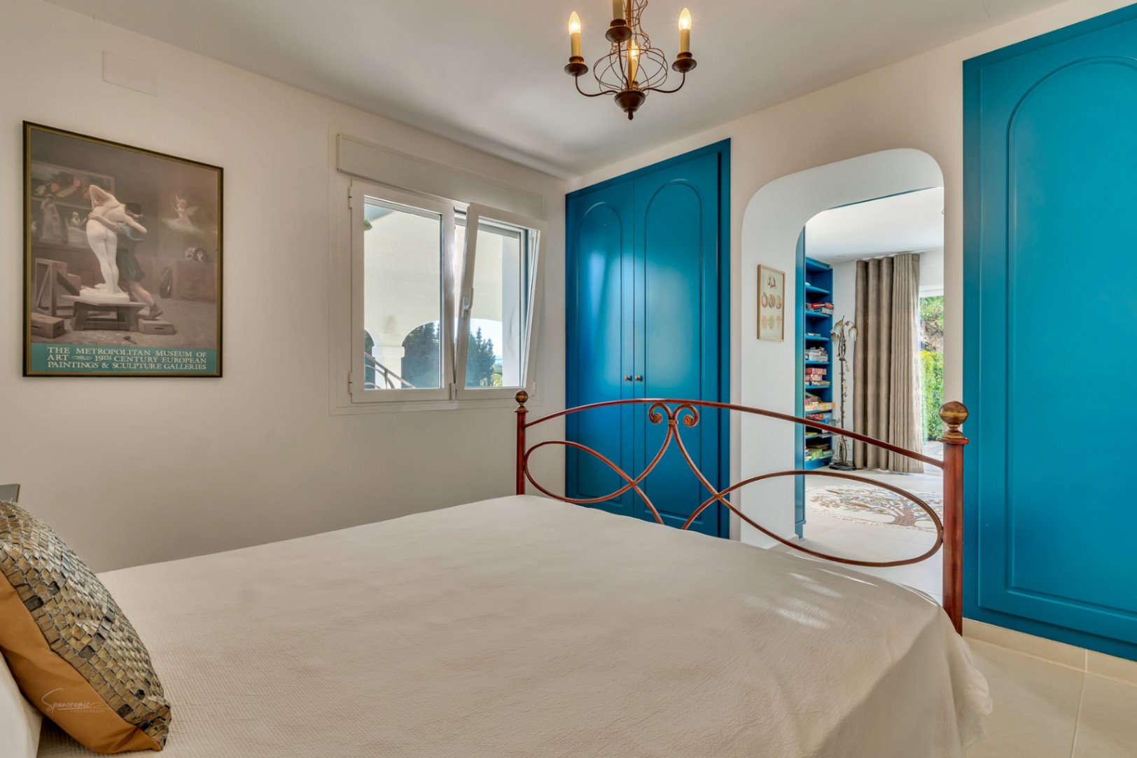Chambre avec placards intégrés bleu vif, lit en fer forgé et passage voûté menant à un espace bureau privé doté d'étagères de rangement.