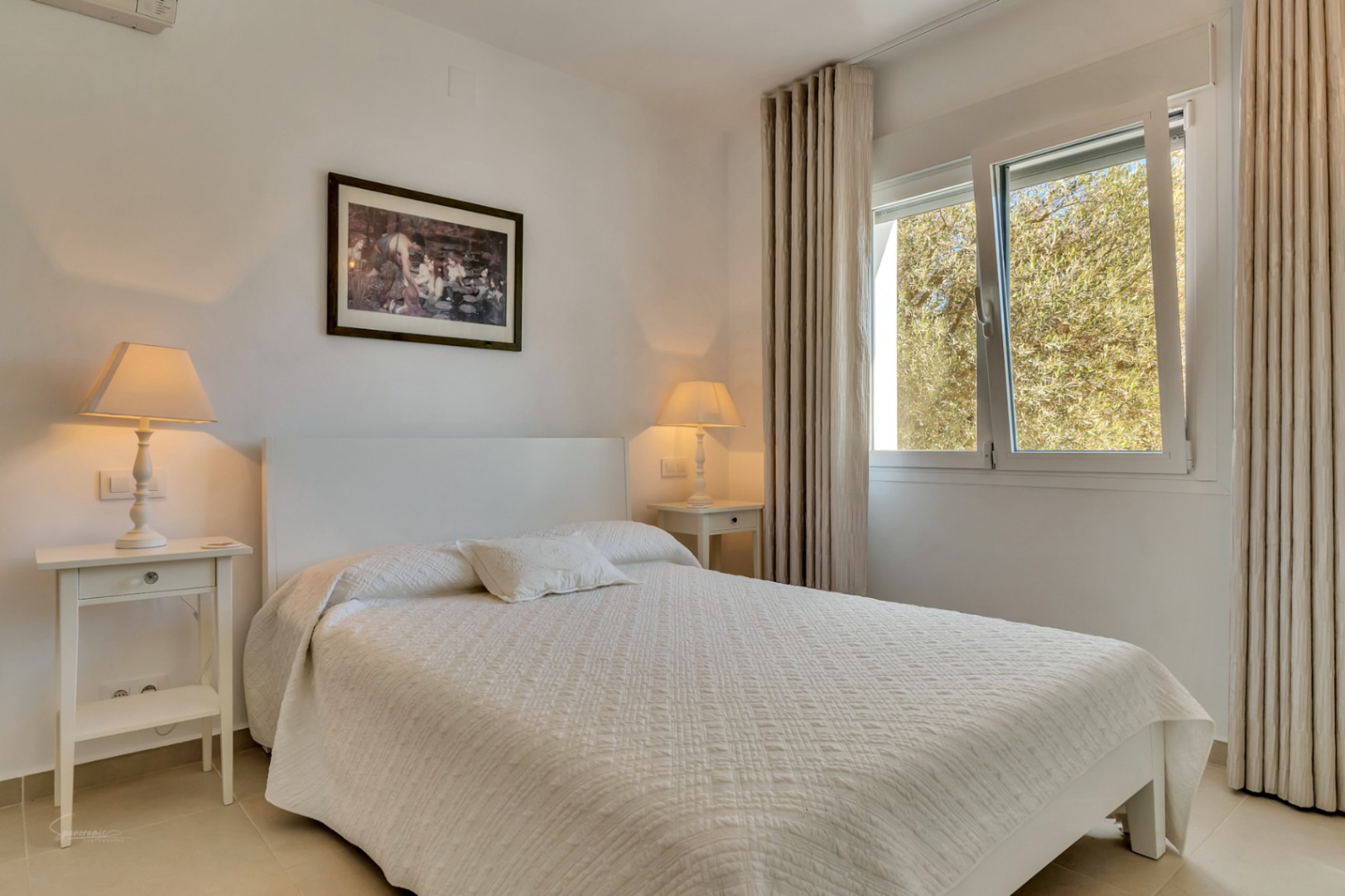 Chambre lumineuse avec lit blanc, tables de chevet assorties et lampes à lumière chaude. La fenêtre avec rideaux neutres offre une vue sur la verdure environnante.