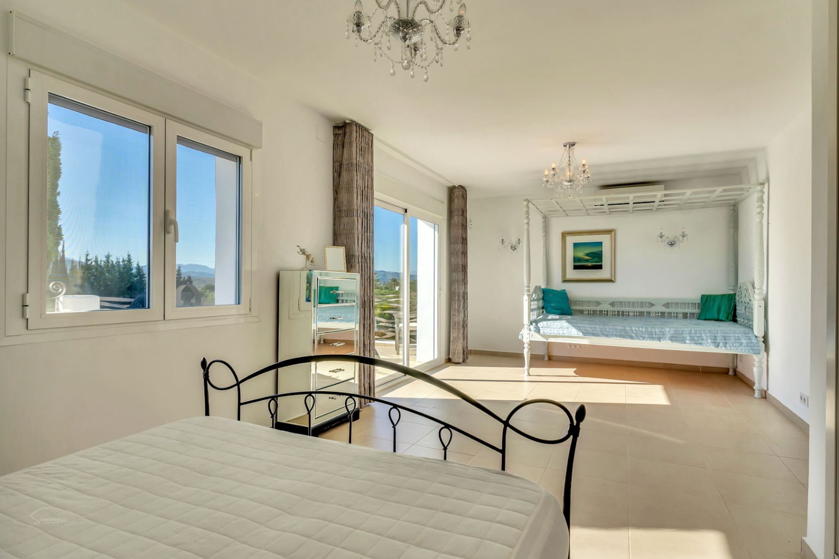 Chambre lumineuse avec lit en fer forgé, commode en miroir et méridienne en bois sculpté sous des lustres en cristal, offrant un accès direct à la terrasse.