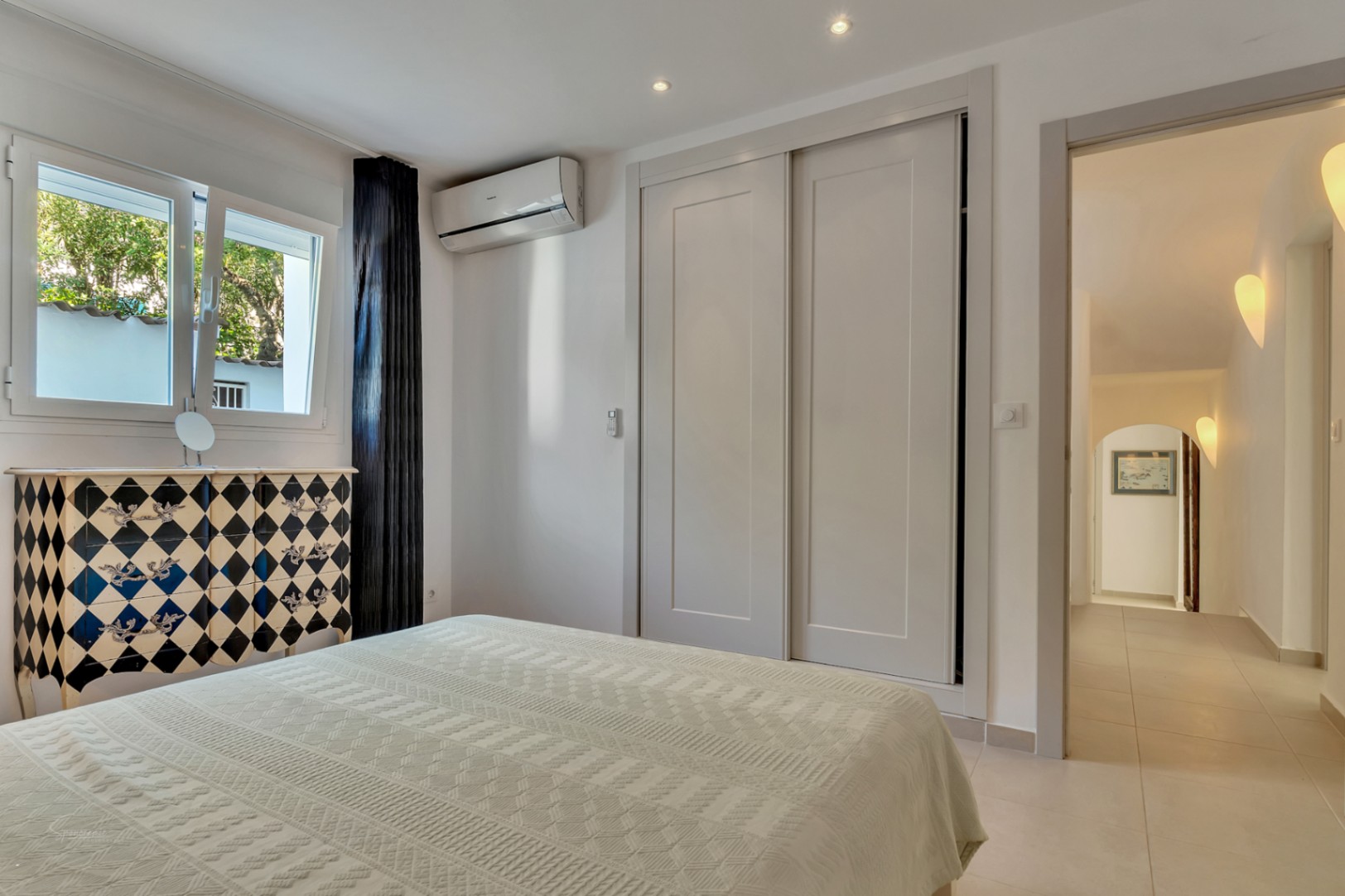 Chambre lumineuse avec placards intégrés, climatisation et commode décorative. La pièce s'ouvre sur un couloir éclairé par des appliques murales élégantes.